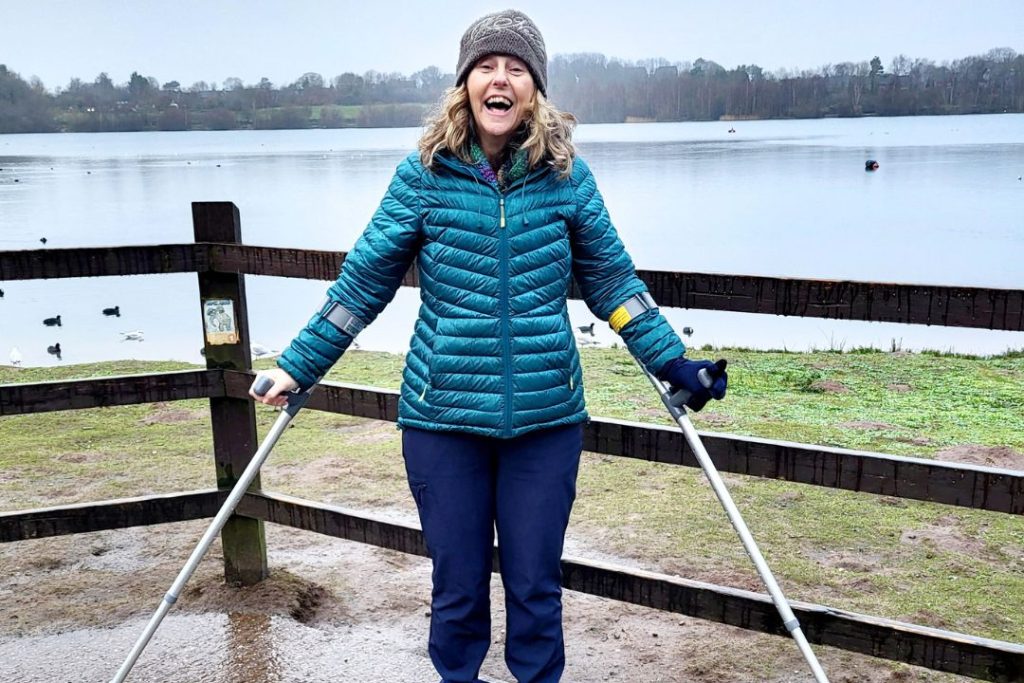 Founder of walking for charity app trundl Hil Mines stands, resting on her crutches, in front of a lake on an autumnal day smiling.