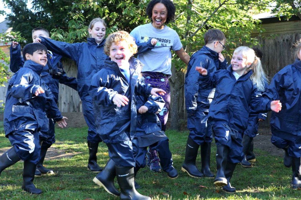 A number of young children in wet weather gear from trundl partner The Outdoor Guide Foundation jump for joy in the sunshine