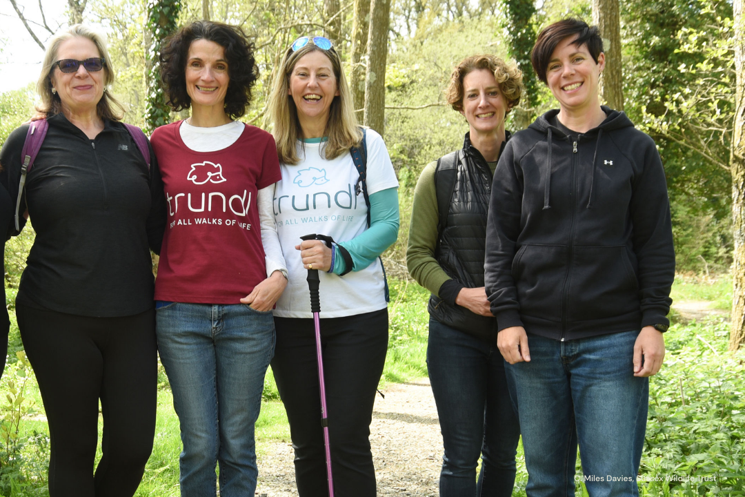 5 women stand together smiling against a wooded background. The 2 founders of the walking for charity app trundl wear bright trundl T-shirts